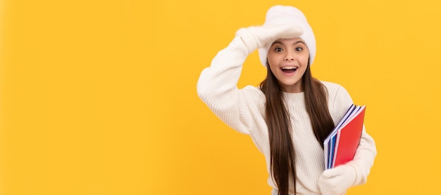 École d'hiver étonnée adolescente ion chapeau et gants d'hiver tenir un cahier sur fond jaune