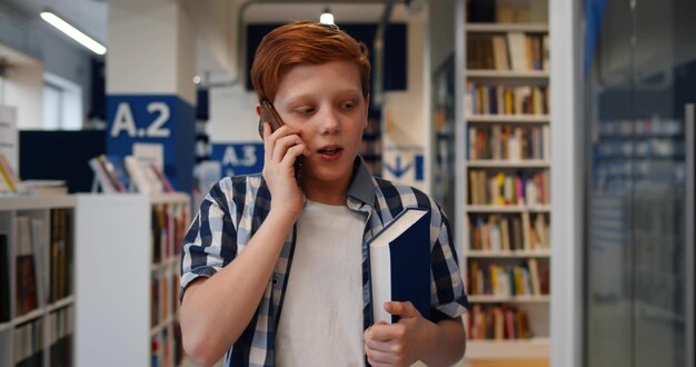 Écolier adolescent marchant dans la bibliothèque de l'école et parlant sur smartphone