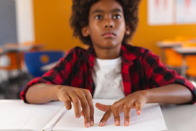 Écolier afro-américain aveugle assis au bureau en classe lisant un livre en braille avec les doigts