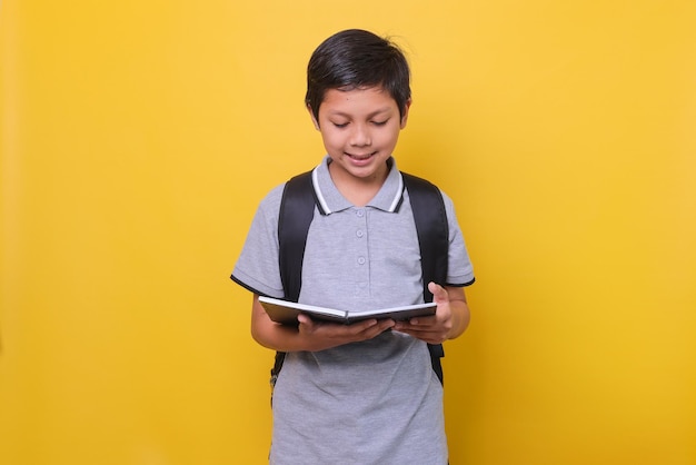 Écolier asiatique dans un style décontracté avec sac à dos noir sourit en lisant un livre isolé sur jaune