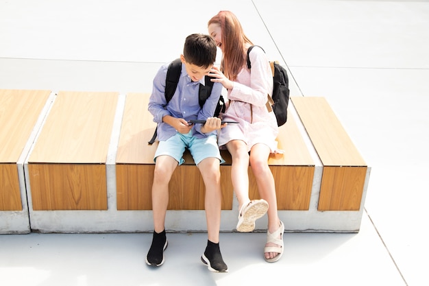 Écolier écolière adolescents rire s'amuser après les cours assis sur un banc en bois dans la cour d'école, tablette d'utilisation de fond en béton, concept d'éducation en ligne, technologies modernes dans la vie des enfants