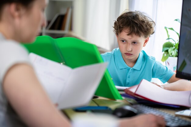 Écolier est assis à table avec sa mère et fait ses devoirs, regardant le manuel