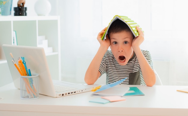 Écolier garçon assis au bureau apprenant caché derrière un cahier d'étude bonne nouvelle note la plus élevée réussir les examens test école, accomplir ses devoirs heureux excité surpris