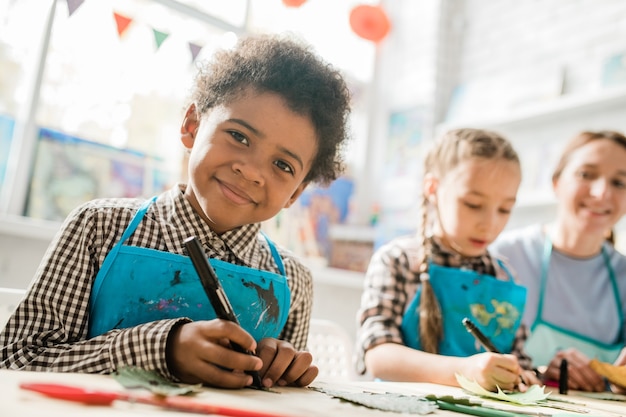 Écolier heureux avec surligneur en vous regardant assis par un bureau à la leçon sur fond de camarade de classe et enseignant