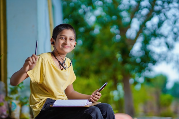 Écolier indien tenant un cours d'apprentissage à distance par téléphone à l'aide d'une application mobile, regardant une leçon en ligne, appel vidéo dans l'application prenant des notes en étudiant à la maison