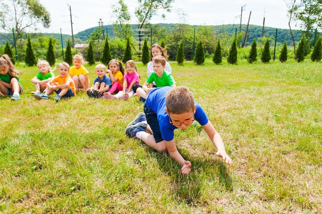 Écolier à lunettes qui croule à genoux sur l'herbe devant ses camarades, assis sur un terrain à l'extérieur dans le parc d'été. Leçons sur l'amélioration des compétences d'acteur pour les enfants d'âge différent.