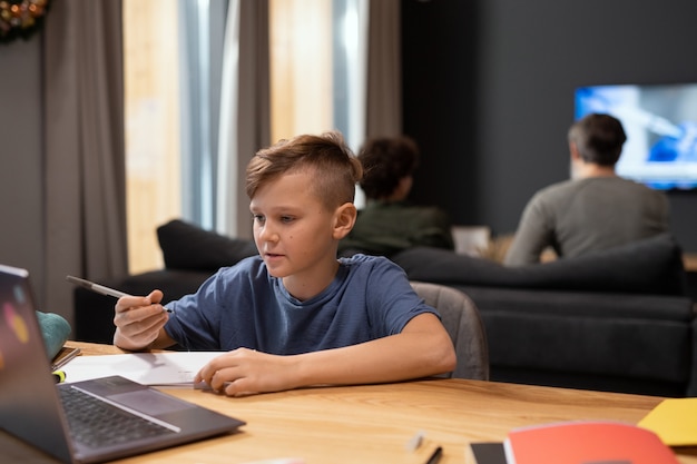 Écolier mignon et intelligent avec un stylo regardant attentivement l'écran de l'ordinateur portable tout en étant assis près d'une table et en écoutant son professeur pendant la leçon en ligne