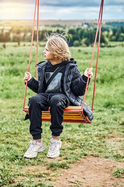 Écolier mignon avec de longs cheveux blonds se promène sur une balançoire regardant de côté par temps nuageux dans la campagne