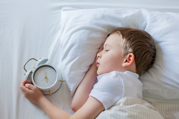 Écolier avec réveil dormant dans son lit. Matin.