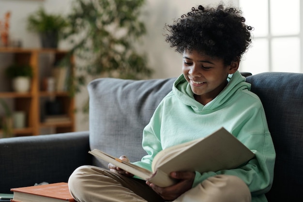 Écolier souriant en tenue décontractée lisant un livre assis sur un canapé