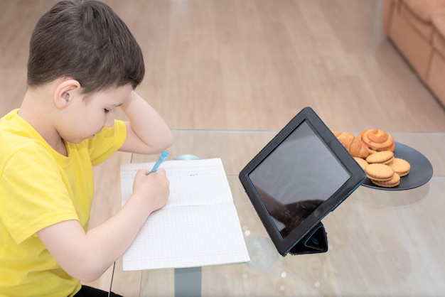 Écolier en t-shirt jaune assis à la table avec tablette numérique étudiant à la maison