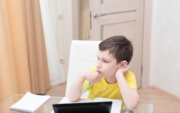 Écolier en t-shirt jaune assis à la table avec tablette numérique étudiant à la maison