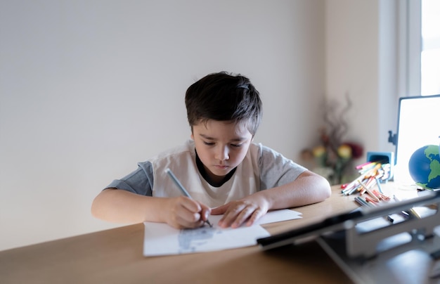 Écolier utilisant un stylo de couleur grise dessinant ou esquissant sur papierPortrait garçon assis sur une table faisant ses devoirs dans le salonL'enfant profite d'activités artistiques et artisanales à la maison le week-endConcept éducatif