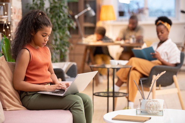 Écolière africaine sérieuse assise sur un canapé dans le salon avec un ordinateur portable sur les genoux et préparant un devoir à domicile contre sa famille