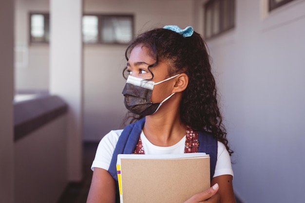 Écolière afro-américaine en masque facial debout dans le couloir de l'école tenant des livres