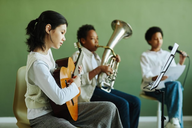 Écolière asiatique mignonne jouant de la guitare acoustique et regardant des notes sur le pupitre tout en étant assis à l'agai