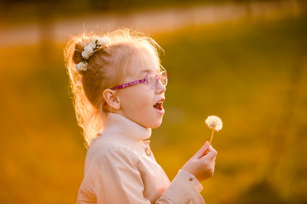 Écolière bouclé dans les lunettes et manteau léger, jouer avec pissenlit dans le parc en automne sur le coucher du soleil