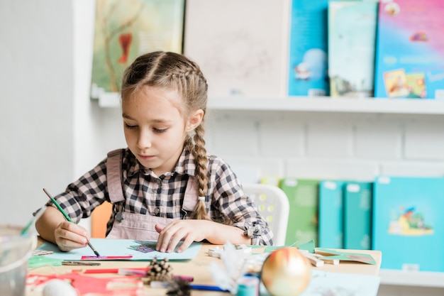 Écolière créative avec pinceau assis par bureau et dessin photo de Noël avec des peintures en classe