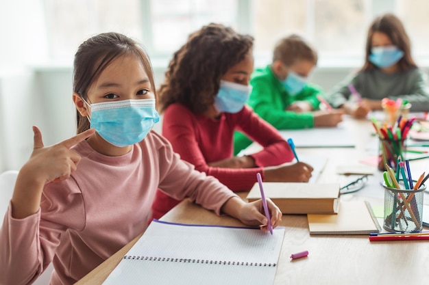 Écolière japonaise portant un masque facial gesticulant les pouces vers le haut dans la salle de classe