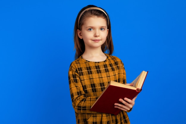 Écolière mignonne attirante sérieuse tenant un livre sur le fond bleu