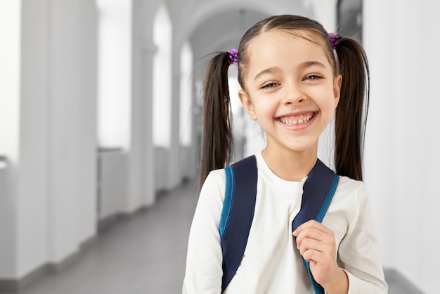 Écolière mignonne, jolie et heureuse avec des cheveux en queue debout dans le long couloir lumineux de l’école primaire souriante