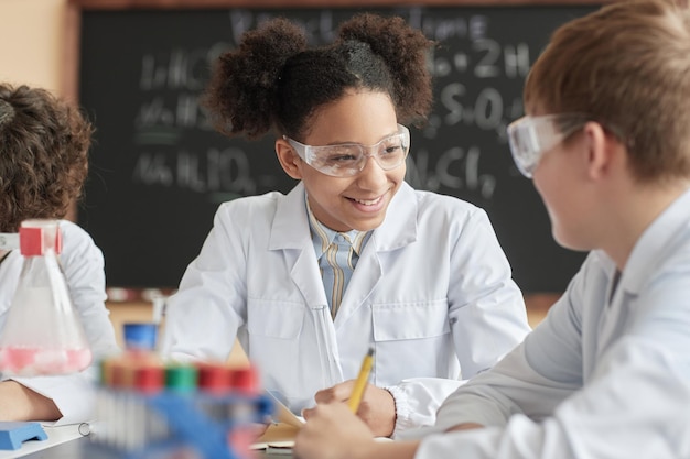 Écolière noire souriante portant une blouse de laboratoire en classe de sciences parlant à un ami