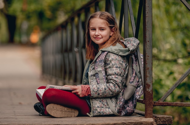 Écolière préadolescente avec sac à dos assis sur le sol et livre de lecture à l'extérieur. Douce élève de sexe féminin étudiant dans le parc d'automne