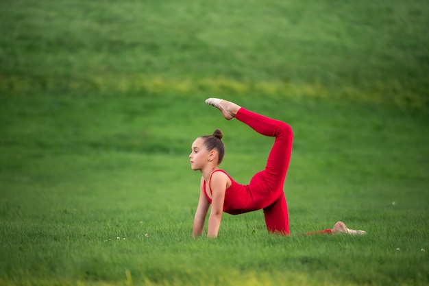 Écolière en salopette rouge vif est engagée dans la gymnastique sur l'herbe, faisant des exercices d'étirement