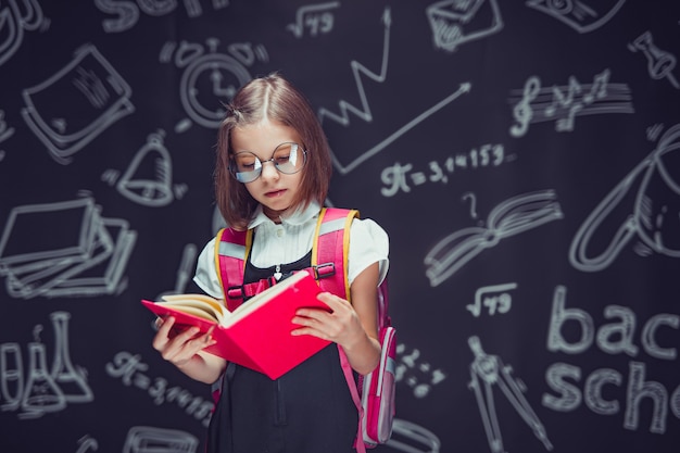 Écolière sérieuse dans des verres se préparant à aller à l'école avec un livre de lecture de sac à dos à l'école