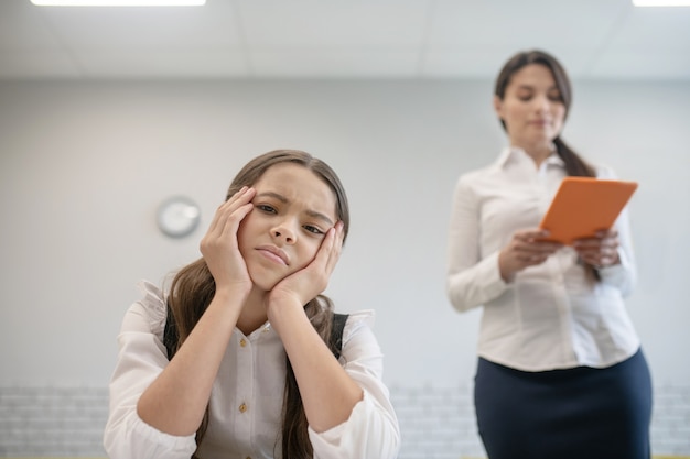 Écolière triste reposant sa tête avec les mains assis au bureau et enseignant avec tablette derrière