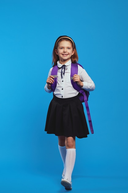 Écolière en uniforme et sac à dos souriant et marchant contre le bleu