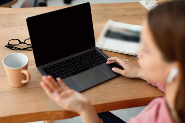 Écolière utilisant un ordinateur portable avec écran vide faisant des gestes tout en parlant à la webcam assis au bureau à la maison ayant un appel vidéo