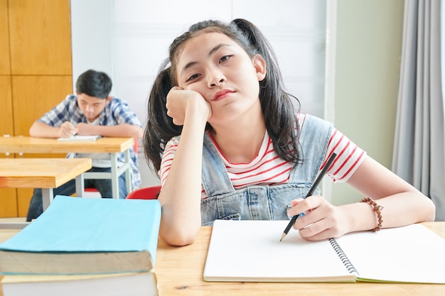 Écolière vietnamienne s'ennuie assis au bureau en classe et écrit dans un cahier