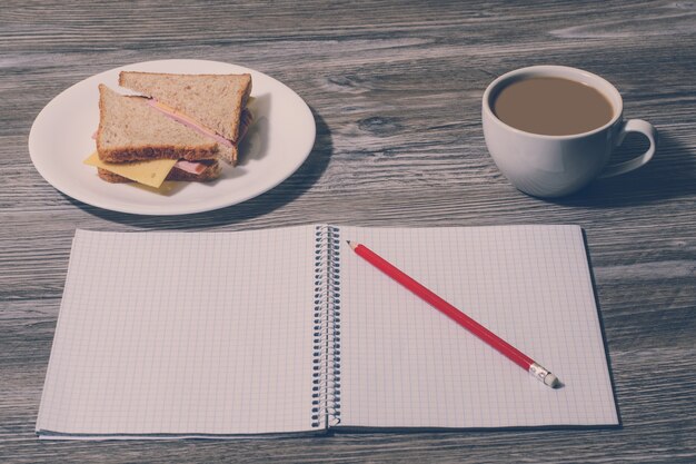 Photo collation au travail. sandwich au fromage savoureux sur une assiette, tasse de café chaud, cahier ouvert avec un crayon sur fond de bois gris. vue de dessus, effet vintage