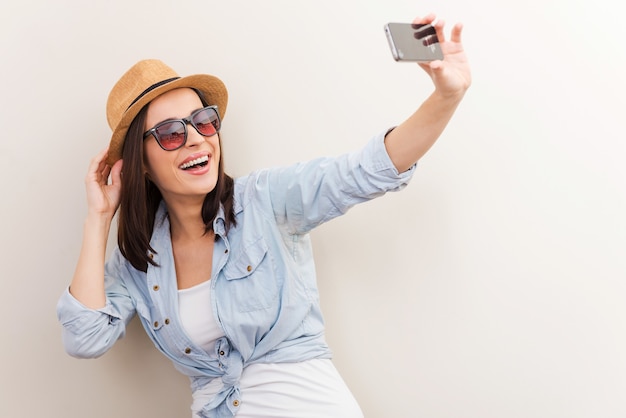 Collecter des souvenirs. Portrait de la belle jeune femme à lunettes ajustant son chapeau tout en faisant du selfie et debout sur fond marron