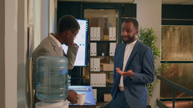 Des collègues afro-américains heureux profitant d'une bonne discussion sur le réservoir d'eau pendant la pause de nuit. Un employé souriant prend une tasse de café, se détend tout en parlant avec un collègue au bureau.