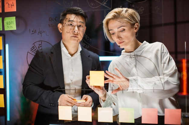 Collègues caucasiens et asiatiques debout au bureau derrière le panneau de verre