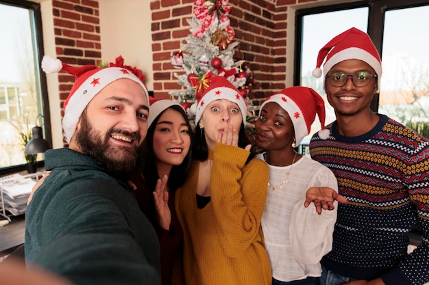 Des collègues divers et souriants portant des chapeaux de Père Noël posent pour un selfie ensemble au bureau avec des décorations de Noël Des collègues joyeux prennent une photo de groupe lors d'une fête d'année nouvelle.
