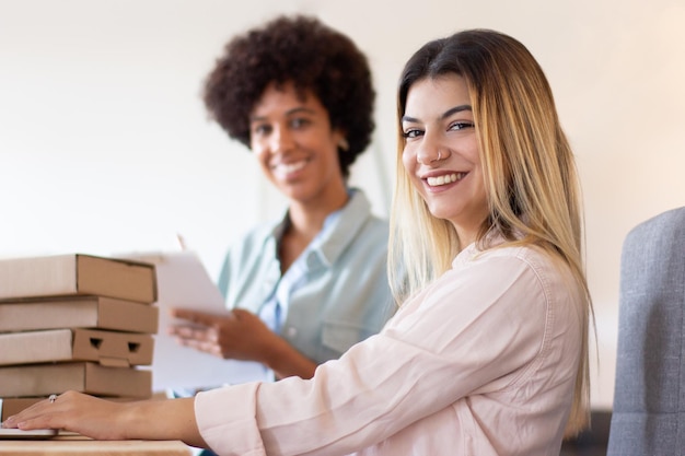 Collègues d'entrepôt travaillant avec des colis. Portrait of smiling young Hispanic woman using laptop while blackwoman examinant les listes de numéros de commande. Concept de démarrage en ligne