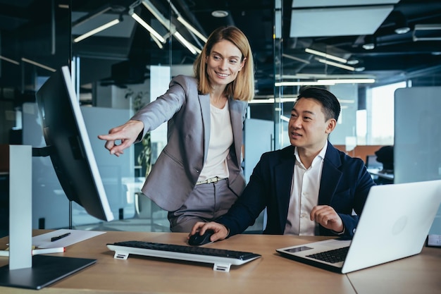 Des collègues heureux discutent de la stratégie du projet en regardant et en pointant sur l'écran du moniteur d'ordinateur portable pc équipe commerciale multiethnique au bureau confiant homme asiatique mature expliquant la jeune femme