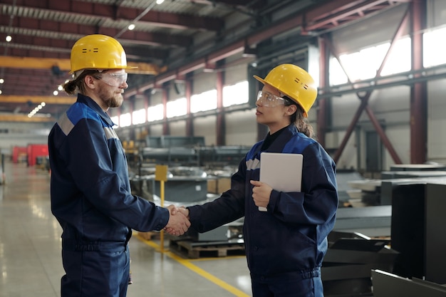 Collègues industriels confiants en vêtements de travail et lunettes de protection faisant une poignée de main en atelier après avoir conclu un accord