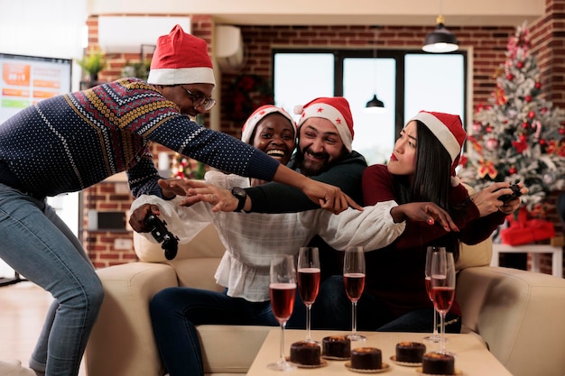 Des collègues joyeux se battent pour le joystick de la console de jeu tout en célébrant Noël dans un bureau décoré. Heureux collègues divers ayant une compétition de jeux vidéo lors de la fête d'entreprise du nouvel an