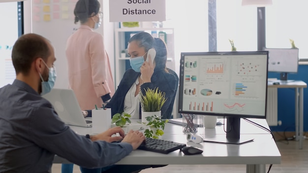 Des collègues avec des masques faciaux assis au bureau dans un nouveau bureau d'entreprise normal travaillant sur un ordinateur portable, parlant au téléphone pendant la pandémie de coronavirus. L'équipe maintient une distance sociale pour éviter les maladies virales