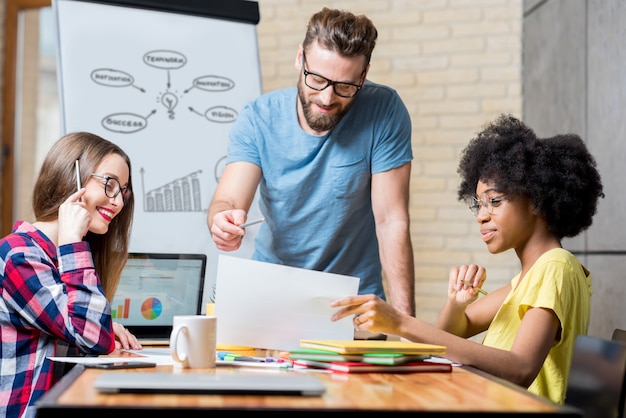 Des collègues multiethniques vêtus avec désinvolture de vêtements colorés travaillant pendant la réunion au bureau avec un ordinateur portable, des documents et un tableau blanc