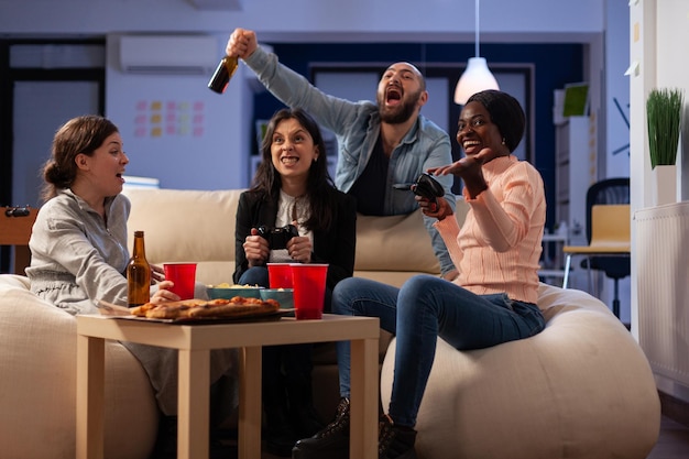 Des collègues de travail heureux célébrant la victoire des jeux vidéo avec des boissons dans le bureau d'affaires après les heures d'ouverture. Collègues s'amusant avec le jeu en ligne, dégustant de la nourriture et des boissons alcoolisées lors de la célébration.