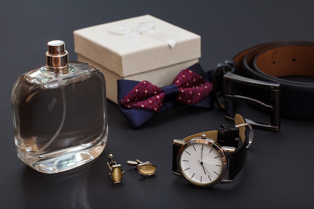 Cologne pour homme, coffret cadeau blanc, noeud papillon, boutons de manchette, montre avec un bracelet en cuir noir et ceinture en cuir avec boucle en métal sur fond noir. Accessoires pour hommes.