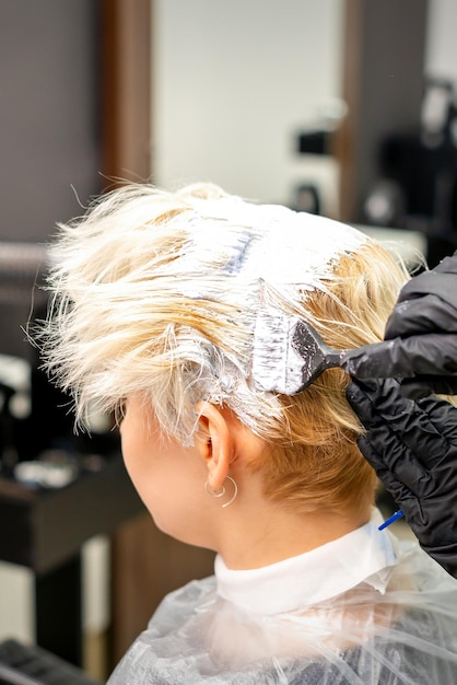 Coloration des cheveux blancs avec teinture pour les cheveux et brosse par les mains du coiffeur pour la jeune femme blonde caucasienne dans un salon de coiffure en gros plan