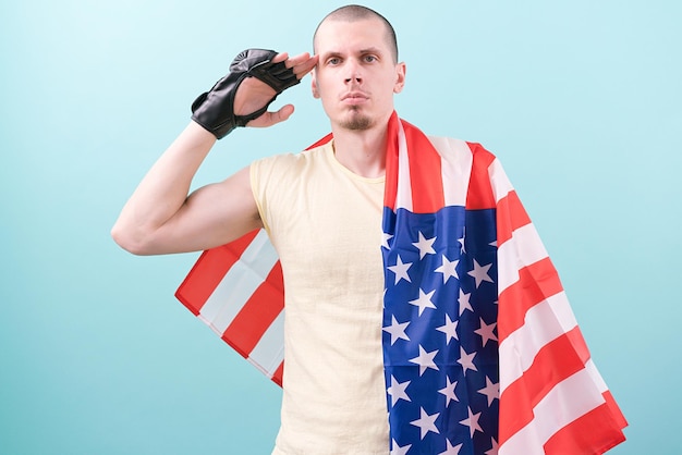 Un combattant caucasien de MMA dans des gants noirs et recouvert d'un drapeau américain tient sa main près de sa tête sur un fond bleu un Force Fighting Respect Military Service Saluting Discipline Duty