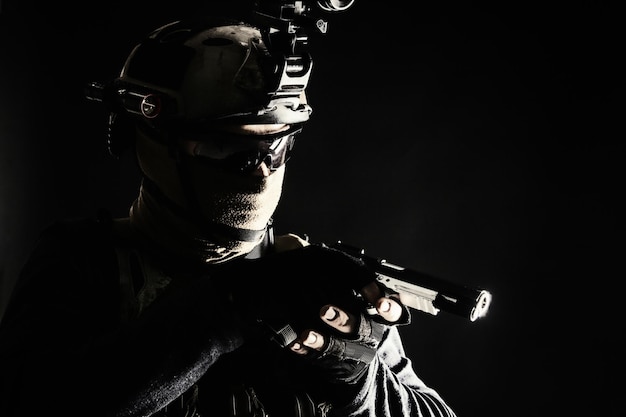Combattant de l'équipe SWAT de la police de l'escouade antiterroriste de l'armée cachant son identité derrière un masque et des lunettes portant un casque avec un dispositif de vision nocturne rampant dans l'obscurité visant un pistolet de service pendant la mission