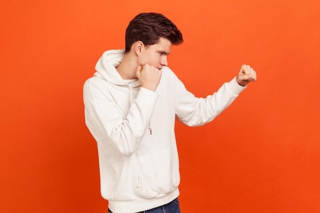 Combattant masculin en sweat à capuche tenant les poings serrés, prêt à attaquer. Jeune homme se défendant des intimidateurs. Cours d'école d'arts martiaux. Studio intérieur tourné isolé sur fond orange